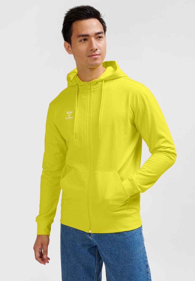 Mand iført en lys gul lynlåstrøje med logo, står med den ene hånd i lommen mod en ensfarvet lys baggrund.