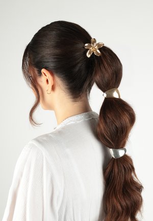 Donna con lunghi capelli castani acconciati in una coda di cavallo a bolle fissata da tre fermagli dorati, che indossa una maglia bianca con trama.