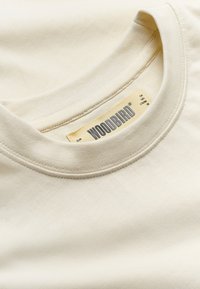 Κοντινό πλάνο του ντεκολτέ ενός εκρού t-shirt που δείχνει μια μπεζ ετικέτα με την επωνυμία "WOODBIRD" και λεπτομέρειες μεγέθους.