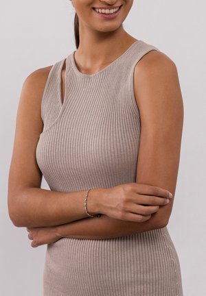 Beige, geribbelde mouwloze jurk met een uitsnede-ontwerp. Het model draagt een delicate zilveren armband en kruis haar armen, waarmee ze de textuur van het kledingstuk benadrukt.