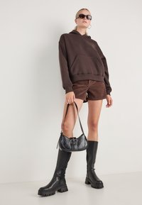 Hanorac călduros maro, pantaloni scurți din suede maro, cizme negre cu toc bloc până la genunchi și o geantă neagră cu detalii tip cataramă.
