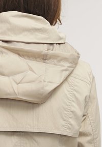 Chaqueta con capucha beige de textura suave y brillante. Presenta una cremallera y detalles cosidos en la parte trasera. Diseño de capucha ligeramente fruncida.