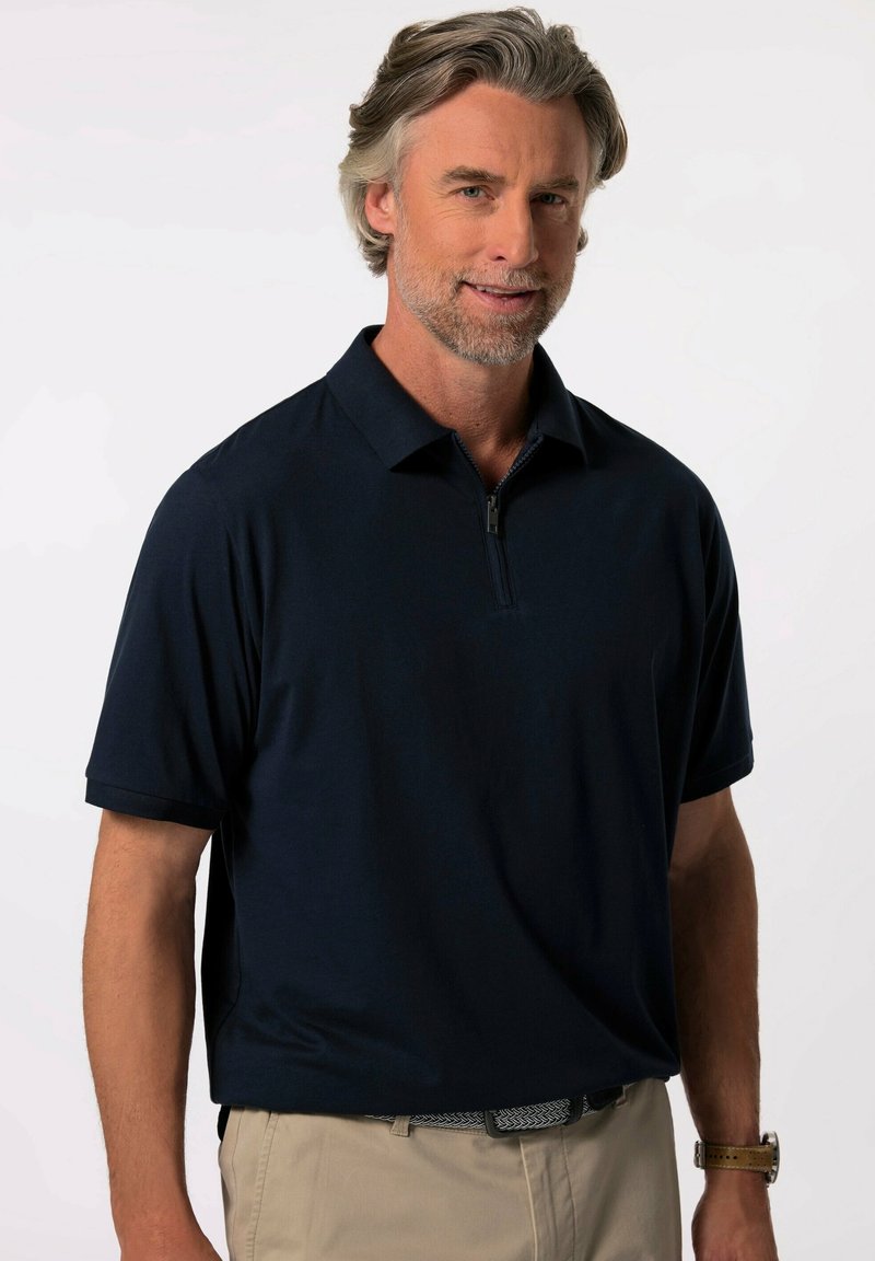 Laivasto sininen polo paita lyhyillä hihoilla ja vetoketjukauluksella, valmistettu sileästä kankaasta; yhdistetty vaaleanvärisiin housuihin ja kuviolliseen vyöhön.