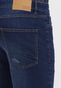 Tmavomodré denimové džínsy s textúrovaným povrchom, s koženou nášivkou označenou "THE DENIM JOURNEY" a zadným vreckom s detailným prešitím.