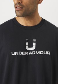 Fekete pamut póló kerek nyakkal, rövid ujjakkal, a elején egy nagy, fehér "UNDER ARMOUR" logóval, amelyen stilizált "U" betű található.