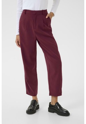 Frau trägt hoch taillierte burgunderrote Hose mit Falten und schwarze Loafers, steht mit einer Hand in der Tasche vor weißem Hintergrund.