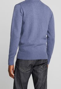 Blå stickad tröja med ribbad nederkant och mudd, med en texturerad yta. Matchad med mörka denimbyxor. Bakvy visas.