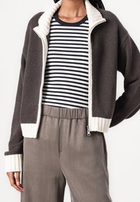 Kurtka z fleece'em, zapinana na zamek, w ciemnoszarym kolorze z kontrastowym kremowym kołnierzem i mankietami. Pod nią pasek w poziome paski, w zestawieniu z szarymi spodniami.