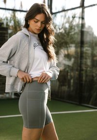 Šedé sportovní šortky s vysokým pasem a bočními kapsami, spojené s bílým tričkem a světle šedou mikinou na zip. Přirozené osvětlení.