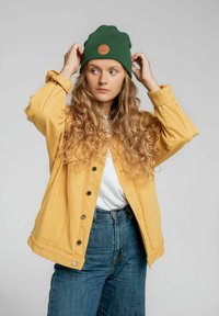 Żółta dżinsowa kurtka z czarnymi guzikami, noszona na białym t-shircie, w połączeniu z niebieskimi spodniami z wysokim stanem i zieloną dzianinową czapką.