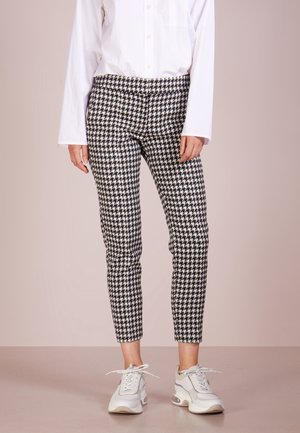 Osoba nosząca czarno-białe dopasowane spodnie w wzór jodełki, białą koszulę z długim rękawem oraz białe trampki, stojąca na jasnym tle.