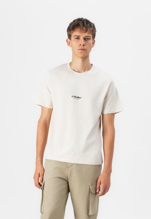 Short-sleeve cream t-shirt made of cotton, featuring a black logo on the chest. Worn with light khaki cargo pants, displaying side pockets.