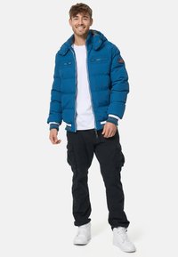 Blue puffer jacket with a zip front, striped cuffs, and dual chest pockets. Paired with black cargo pants and white sneakers.