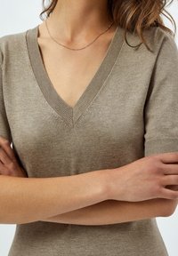 Pull beige à manches courtes avec col en V, comprenant des poignets et un col côtelés, confectionné dans un matériau tricoté doux avec une texture subtile.