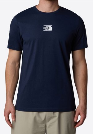 Homme portant un t-shirt bleu marine à manches courtes avec le logo "The North Face" sur la poitrine et un pantalon beige, debout devant un fond uni.