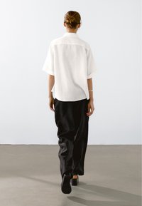 Witte linnen korte mouwen shirt gecombineerd met zwarte losse broek. De persoon draagt schoenen met blokhak en staat met de rug naar de kijker toe.