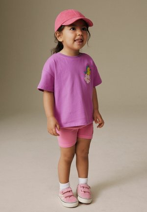 Kisgyermek rózsaszín sapkában, lila pólóban hal motívummal, rózsaszín rövidnadrágban, fehér zokniban és rózsaszín tornacipőben, állva és felfelé nézve.