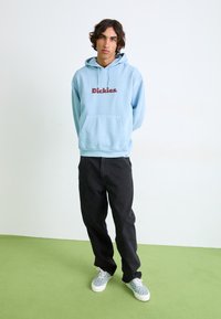 Sky blue hoodie with a red "Dickies" logo, front pocket, paired with black jeans and patterned sneakers on a green floor.