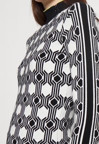 Jersey de patrón en blanco y negro con diseños geométricos. Cuello alto con un detalle en amarillo. Textura de punto y forma ajustada.