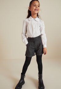 Witte colbert met knopen, grijze hoge shorts met plooien en twee knopen, zwarte legging en zwarte enkellaarsjes.