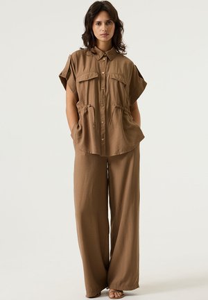 Camisa marrón de manga corta con dos bolsillos delanteros, combinada con pantalones marrones de corte ancho. La tela parece suave y la silueta relajada.