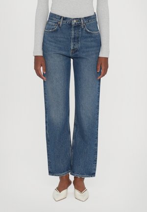 Personne portant un jean bleu taille haute, coupe droite, associé à des talons blancs à bouts pointus et un haut gris clair à manches longues, sur un fond uni.