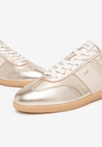 Baskets dorées métalliques avec une texture lisse, ornées de lacets et d'accents blancs. Conçues avec un profil bas et une semelle en caoutchouc beige.