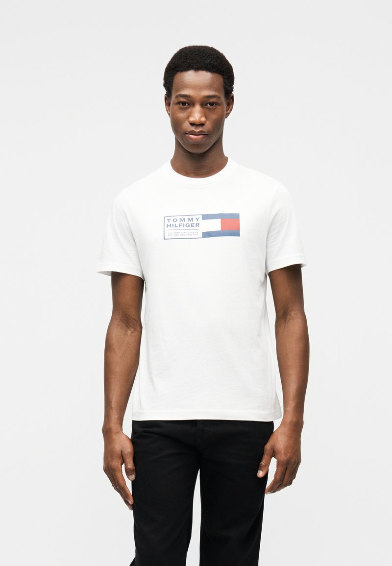 Νέος άνδρας που φοράει λευκό T-shirt Tommy Hilfiger με μπλε, λευκό και κόκκινο λογότυπο, σε συνδυασμό με μαύρο παντελόνι, στέκεται μπροστά σε απλό φόντο.