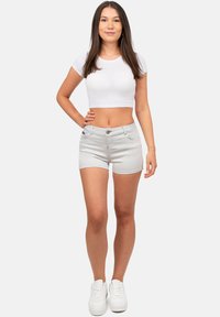 Jeune femme se tenant avec une main sur la hanche, portant un haut court blanc, un short gris clair et des baskets blanches sur un fond uni.