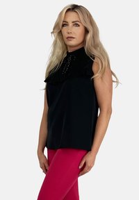 Czarny top bez rękawów z falbaną na kołnierzu i ozdobnymi detalami przy dekolcie, w połączeniu z dopasowanymi różowymi spodniami.