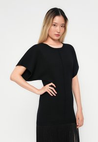 Svart strikket topp med rund halslinning og korte, vide ermer. Designet har en sømløs finish med et frynsekantdetalj.