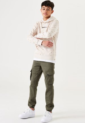 Felpa beige con motivo splatter e scritta nera, abbinata a pantaloni cargo olive con più tasche e sneakers bianche.
