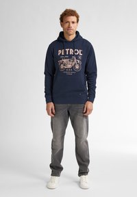 Navyblauwe hoodie met de opdruk "PETROL" en een motorontwerp in lichtroze. G gedragen met grijze jeans en witte sneakers.