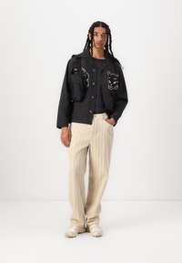 Black jacket with embroidered cat design, gray shirt, beige striped pants, and cream sneakers. Black bag attached to the jacket's shoulder.
