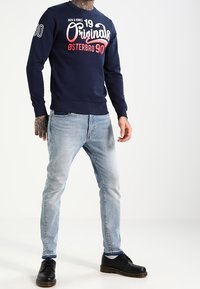 Námorníková modrá mikina s bílým a červeným grafickým textem, spárovaná se světlými modrými džínami a černými botami. Obsahuje žebrované manžety a lem.