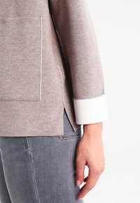 Béžový pletený svetr s přední kapsou, bílými manžetami a bočními rozparky. Nosený s šedými džíny, které ukazují hladkou texturu a uvolněný střih.