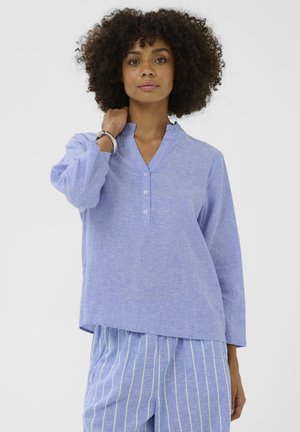 Žena s kučeravými vlasmi oblečená v svetlomodrej košeli na gombíky a modrých pruhovaných nohaviciach pózuje s jednou rukou pri krku na bielom pozadí.