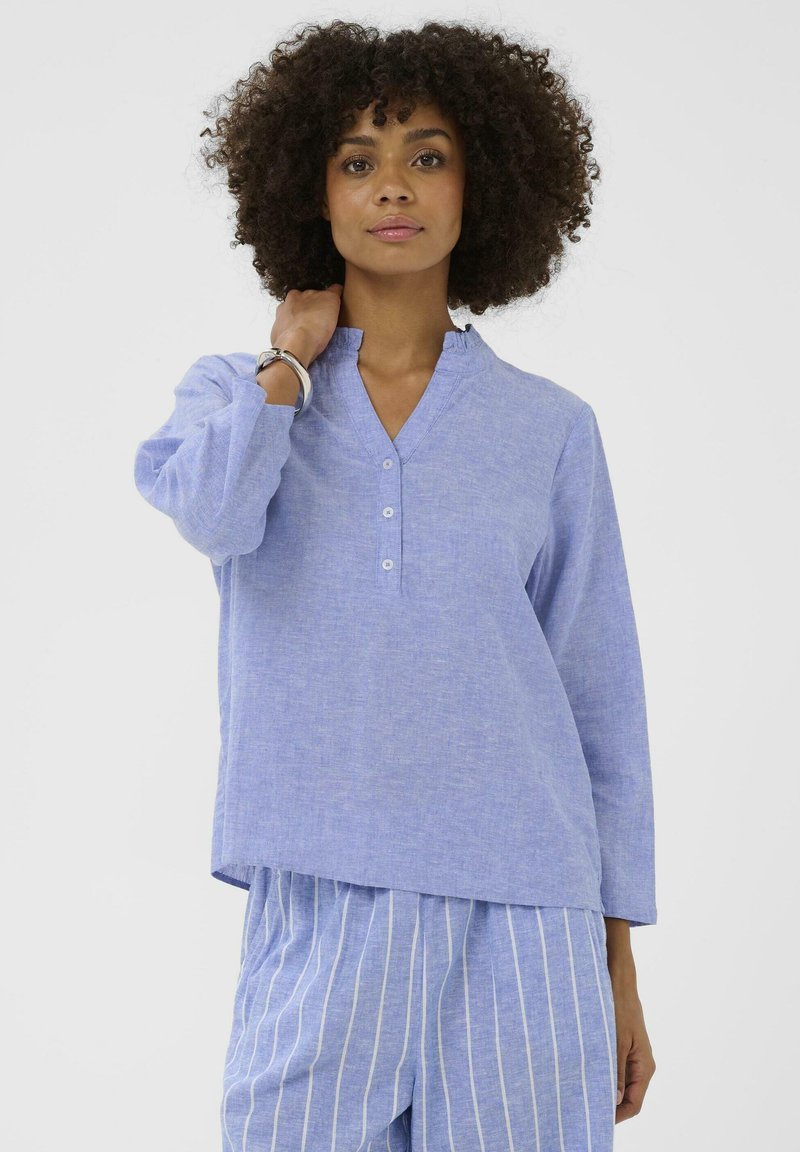 Žena kovrčave kose u svijetloploptoj košulji s dugmićima i plavim hlačama s prugama, pozira s jednom rukom blizu vrata na bijeloj pozadini.