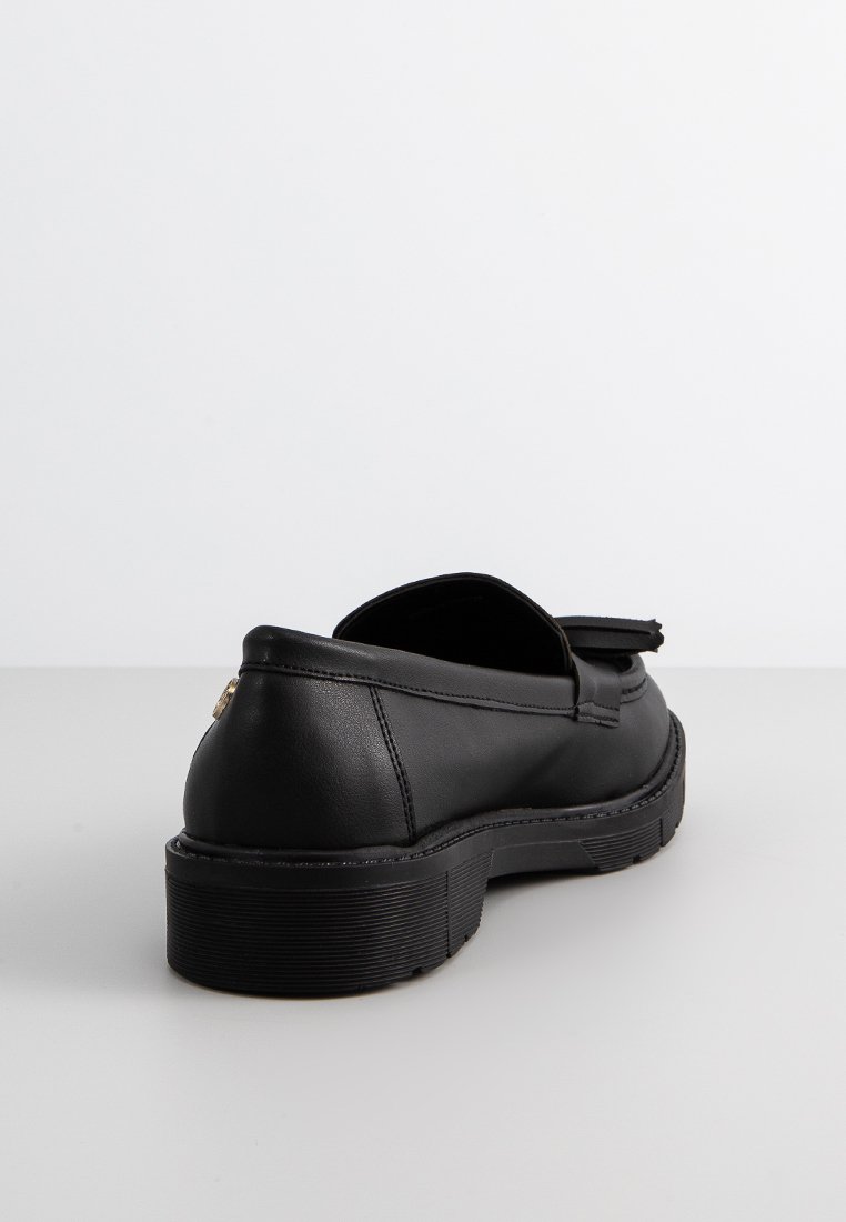 Mocassino in pelle nera con suola spessa e scanalata e dettaglio con nappine, visto da dietro su una superficie bianca uniforme.