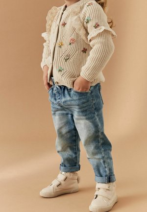 Enfant portant un pull en laine crème avec broderie florale, jean bleu retroussé et baskets crème à scratch, mains dans les poches.
