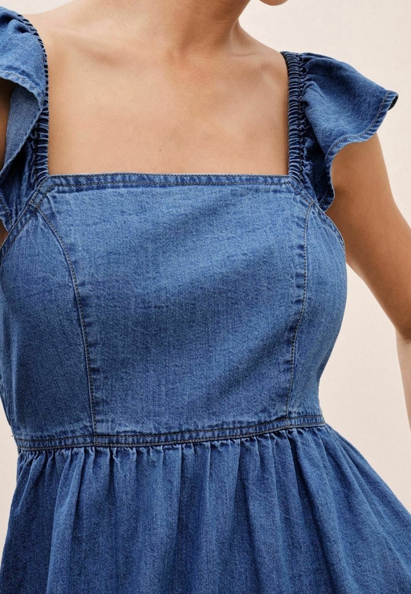 Gros plan sur une personne portant une robe en jean bleue sans manches avec des bretelles volantées aux épaules et un décolleté carré.