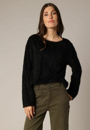 Vrouw met lang bruin haar, gekleed in een zwarte fuzzy trui en olijfgroene broek, staande met één hand op de heup tegen een effen achtergrond.