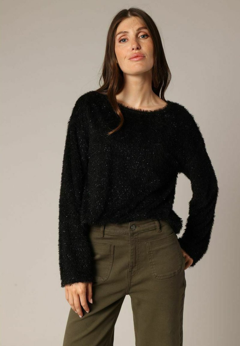 Vrouw met lang bruin haar, gekleed in een zwarte fuzzy trui en olijfgroene broek, staande met één hand op de heup tegen een effen achtergrond.