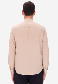 Camicia a maniche lunghe di colore beige chiaro, realizzata in tessuto strutturato. Presenta un colletto con bottoni e un orlo arrotondato con polsini abbottonati.