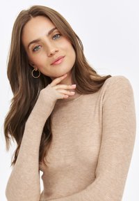 Beżowy sweter z prążkowanej dzianiny z długimi rękawami i otworami na kciuki. Modelka prezentuje miękką teksturę i dopasowany krój, z luźnymi falami we włosach i kolczykami w kształcie kół.
