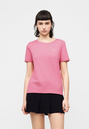 Νεαρή γυναίκα με σκουρόχρωμα μαλλιά φοράει ένα ροζ μπλουζάκι με δύο μικρές περιγραμμμένες καρδιές και μια μαύρη πλισέ φούστα, στέκεται μπροστά σε λευκό φόντο.