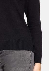Pull en maille noire avec ourlet et poignets côtelés, présentant une texture lisse. Associé à un jean gris ajusté légèrement brillant.