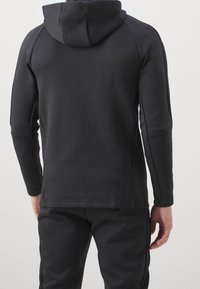 Sweat à capuche noir au design épuré, manches longues et tissu lisse et texturé. Comprend des manches raglan et des coutures discrètes.