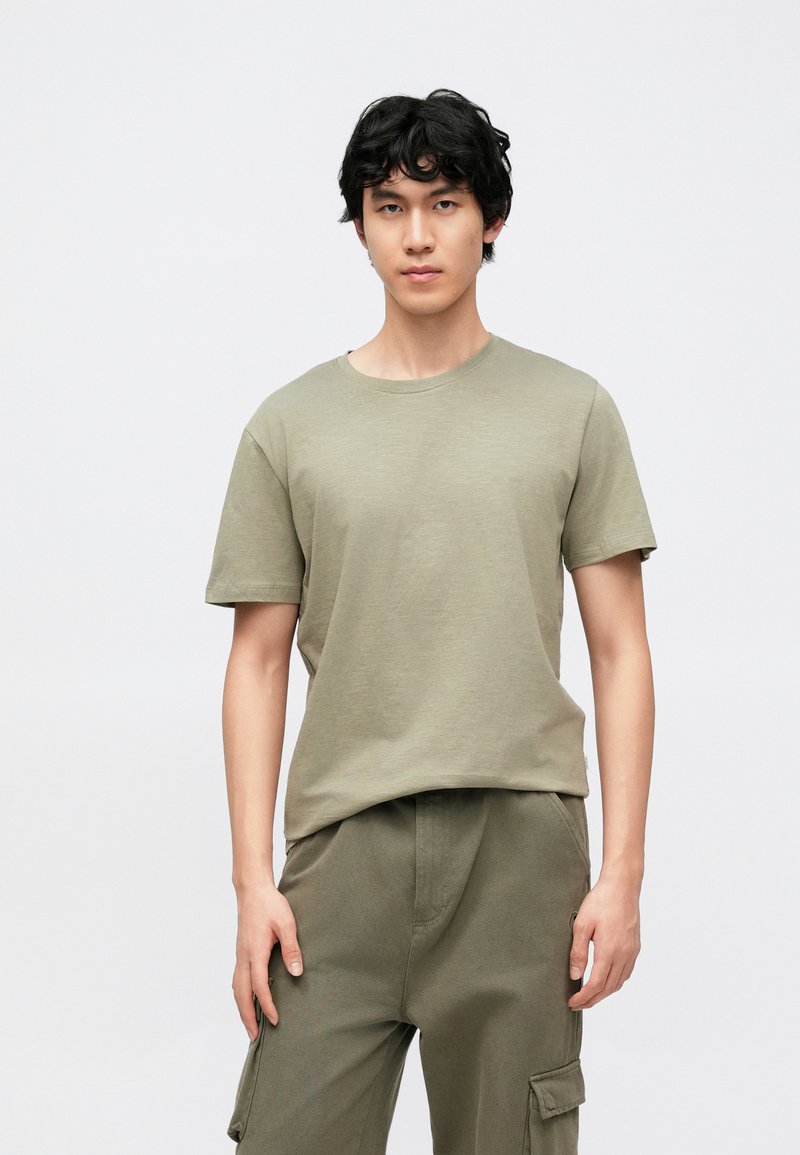 Καραμελλί πράσινη κοντομάνικη μπλούζα από μαλακό ύφασμα, με στρογγυλή λαιμόκοψη και άνετη εφαρμογή, συνδυασμένη με αντίστοιχο παντελόνι cargo.