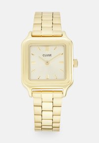 Montre carrée dorée avec un bracelet en métal jaune doré. Le cadran de couleur crème présente des index dorés, le nom de la marque « CLUSE » et une couronne.